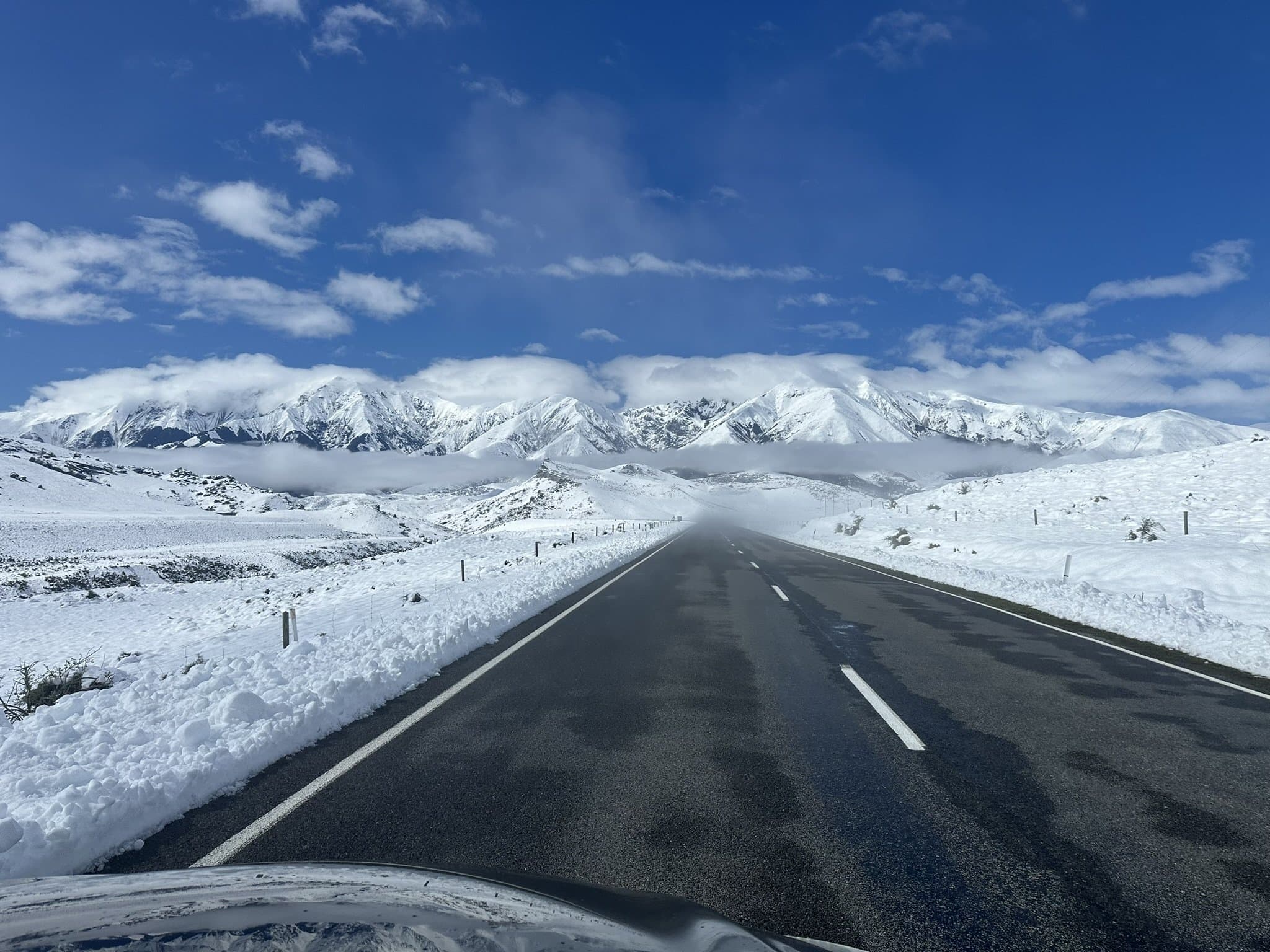 A sealed highway runs dead straight across fresh snow toward a wall of snow-capped mountains in the Canterbury high country